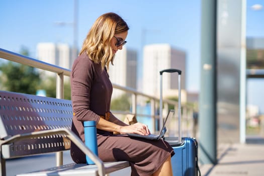 Female manager in stylish dress and sunglasses sitting on bench near suitcase and thermos and browsing netbook on station during business trip in city