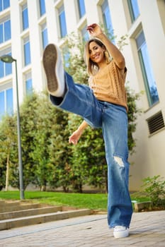 Full body happy adult woman in stylish clothes raising arms and leg and looking at camera with smile while dancing on pavement outside contemporary building