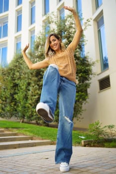 Full body happy adult female in stylish clothes raising arms and leg and looking at camera with smile while dancing on pavement outside contemporary building