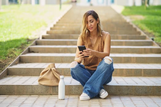 Full body adult female in stylish clothes sitting on stairs near bag and bottle and browsing social media on cellphone on street
