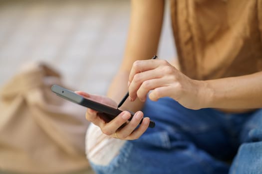 From above soft focus of anonymous female touching screen of cellphone with stylus while sitting on ground on city street in daytime