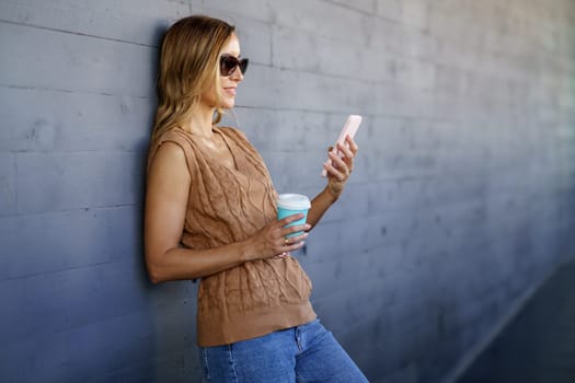 Glad female in stylish clothes and sunglasses, with cup of coffee smiling and browsing social media on cellphone while leaning on gray wall on city street