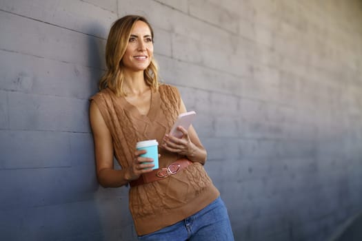 Middle-aged woman taking a coffee break near her office. Caucasian female using a paper cup and carrying her smartphone in her hand.