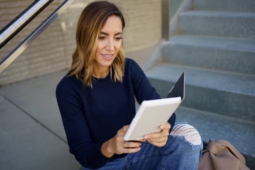 High angle of happy female in stylish jeans and sweater smiling and reading interesting e book while sitting on steps outside modern building