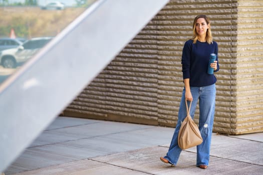 Full body female in stylish jeans and sweater with bag and thermos looking away while standing on pavement outside modern building