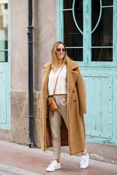 Full body of young female in stylish brown coat sneakers and sunglasses standing on street near building