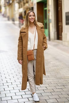 Full body of young blond haired female in trendy brown coat and white sneakers walking on paved street and looking away