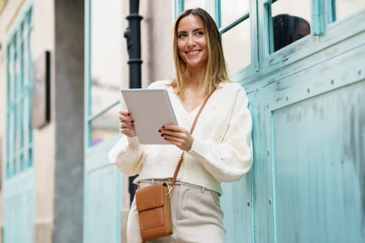 Cheerful blond haired female in smart casual clothes standing on city street while using tablet surfing internet