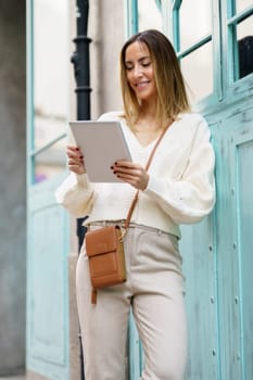 Smiling young woman in smart casual clothes with brown bag standing on city street while using tablet surfing social media