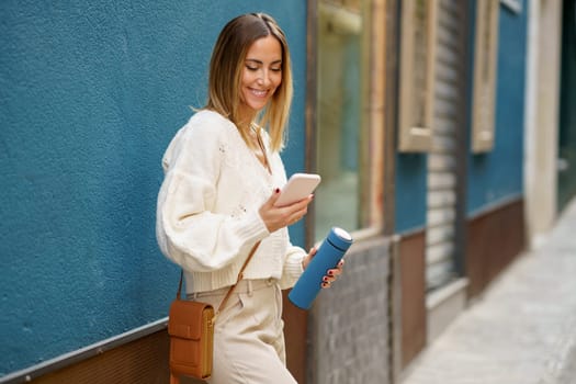 Cheerful young female in smart casual wear standing near blue wall while browsing mobile phone and messaging