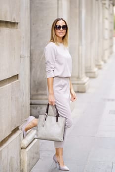 Full body of confident stylish woman gray outfit and sunglasses standing near old building with raised leg and holding handbag at city street