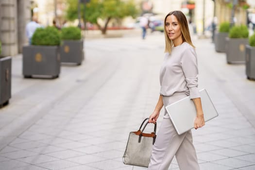 Side view of confident adult female manager in stylish outfit with blond hair looking at camera, while walking on paved street with laptop and bag in hands