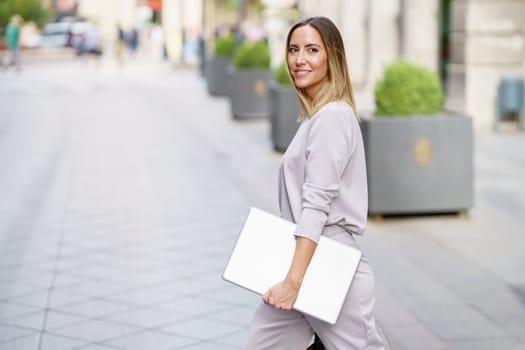 Side view of cheerful adult businesswoman with long blond hair in stylish outfit smiling, and looking away while strolling on paved street with laptop in hand before work