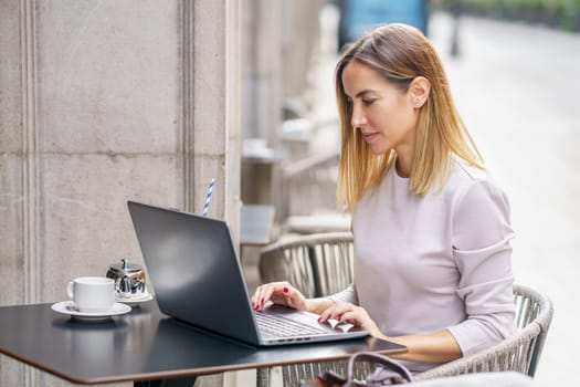 Side view of concentrated adult female freelancer with blond hair in casual clothes reading information on laptop while sitting in street cafe and working remotely