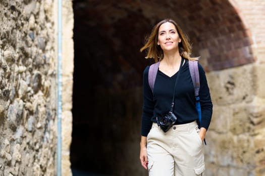 Delighted young female tourist in casual clothes with backpack and photo camera hanging on neck smiling while admiring aged buildings during sightseeing trip