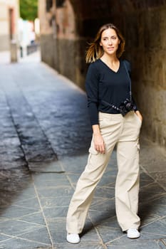 Full body of confident young woman with brown hair and photo camera, in casual clothes standing in stone archway with hand in pocket and looking away in old town