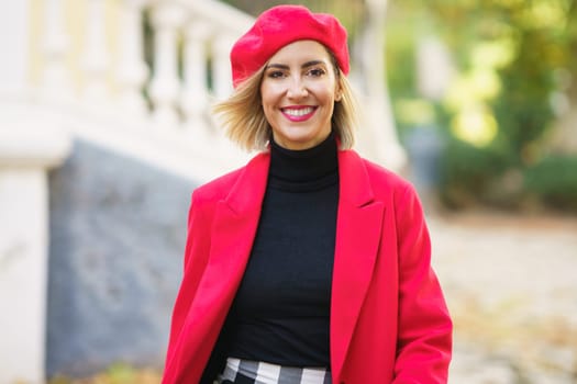 Positive female in trendy red outfit looking at camera with flying hair while strolling in street of city against blurred background