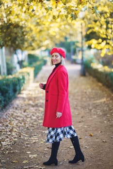 Full body side view of content female in red outfit with beret standing on path with fallen leaves in park on autumn day