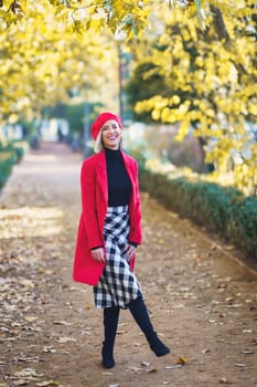 Full body of positive female in stylish outfit looking at camera while standing on pathway in autumn park with trees