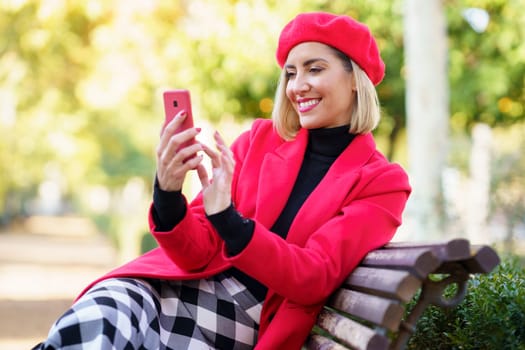 Smiling female in stylish red outfit scrolling social media on modern cellphone while sitting on bench in autumn park on blurred background