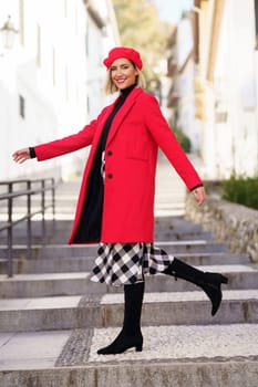 Side view of positive young female in trendy outfit and beret walking on stone street stairs and looking at camera