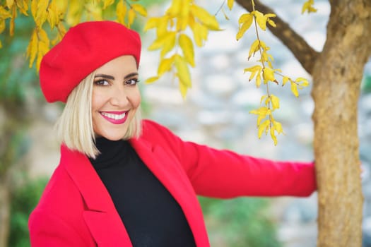 Self assured young content lady, with blond hair and makeup in stylish outfit and beret smiling brightly and looking at camera while touching tree trunk in autumn park