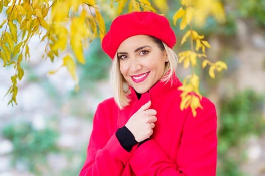 Self assured young blonde with makeup, in stylish red beret adjusting collar of bright coat and smiling while standing near tree in autumn park on sunny day