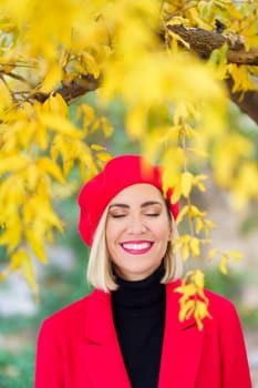 Happy female in bright red beret and coat standing with closed eyes under tree with yellow leaves in park on autumn day against blurred background