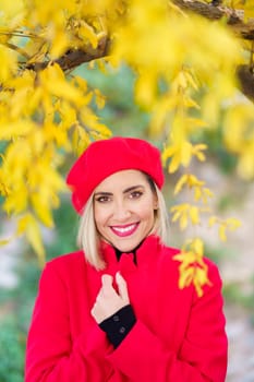 Happy young lady with blond hair and red lips, in bright coat and beret adjusting collar and smiling at camera while standing under yellow tree on sunny autumn day