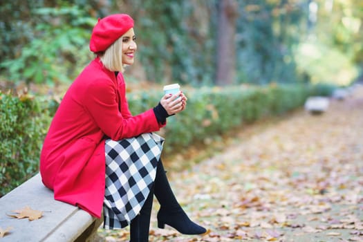 Side view of positive female wearing red beret and coat sitting with legs crossed and having hot takeaway coffee on bench in autumn park