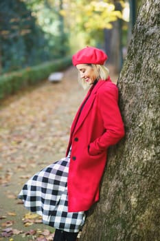 Side view of female standing and hands in pockets of red coat and leaning on tree trunk in autumn park covered with fallen leaves