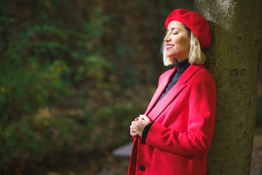 Side view of stylish female wearing red beret and jacket leaning on tree trunk and closing eyes from sunlight in park