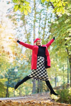 Full body of positive female in bright coat and checkered skirt looking at camera with spread arms while jumping in park on autumn day