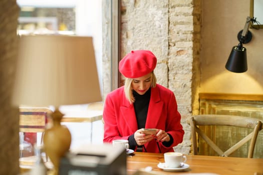 Attractive female in red outfit text messaging on cellphone while sitting at table with cup of coffee in light cafeteria