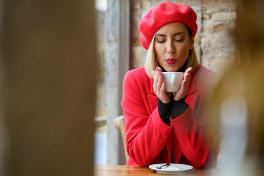 Attractive female in red beret blowing on cup of hot drink in hands while sitting at table in light cafeteria