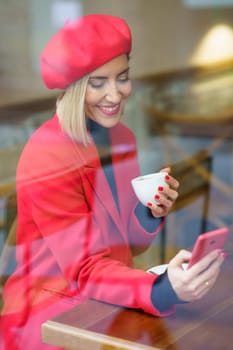 Through window of positive female in red coat and beret text messaging on cellphone while sitting with cup of coffee in cafeteria