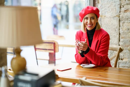 Smiling female in red beret looking at camera with smile while sitting near window with cup of coffee in light cafeteria