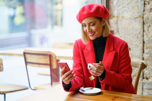 Cheerful female in stylish red outfit scrolling social media on cellphone while sitting at table with cup of tea in cafeteria