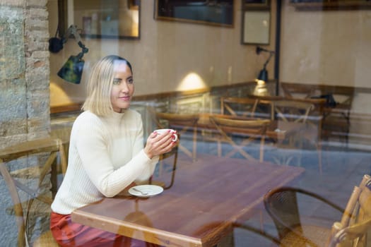 Through glass of dreamy female with cup of hot coffee in hands sitting at wooden table in light modern cafeteria