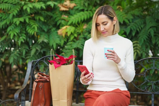 Concentrated female with takeaway coffee surfing cellphone while sitting on bench near paper bag with colorful flowers on street with green plants