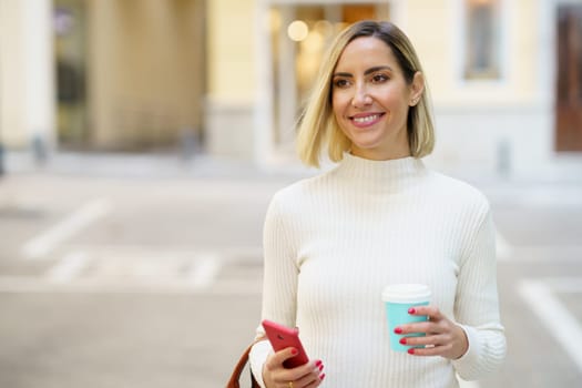 Charming female with disposable cup of takeaway drink and cellphone standing on city street and looking away