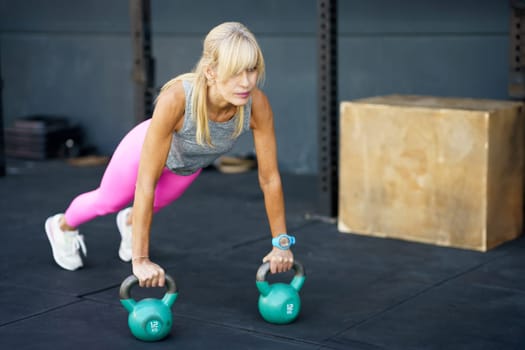 Fit strong blonde hair in activewear doing push ups exercise with heavy kettlebells during functional fitness workout at modern gym against blurred background