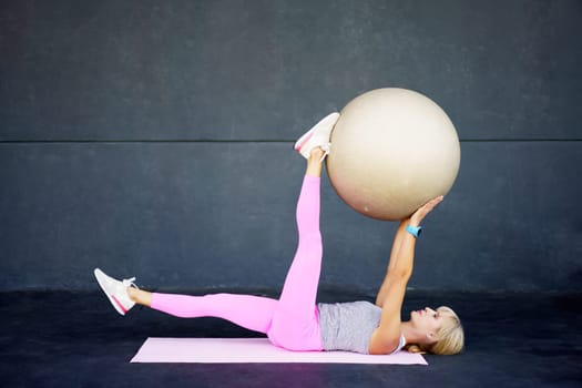 Side view of flexible fit female stretching leg, and doing exercise with fitness ball while lying on mat in modern gym against gray wall during workout
