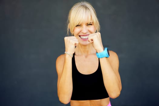 Smiling blonde hair in activewear with blue smartwatch and looking at camera standing against gray background while practices martial art punch