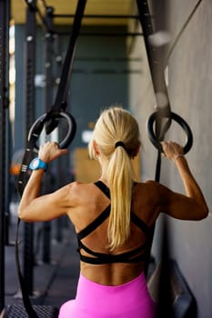 Back view of muscular young fit female with ponytail in activewear exercise on gymnastic rings during fitness training at modern sport club against blurred background