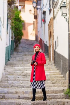 Full body of glad female photographer in classic coat with modern photo camera in hands looking at camera while standing on stairs near buildings