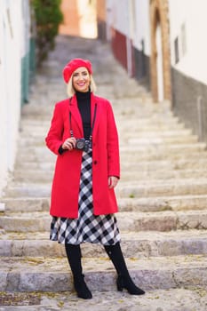 Full body of self assured young blondie, with photo camera on neck in stylish red coat and beret smiling happily while standing on stone steps between aged buildings in town