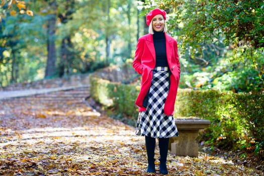 Full body of positive female wearing red coat and beret with checkered skirt standing on fallen leaves in autumn park and smiling happily