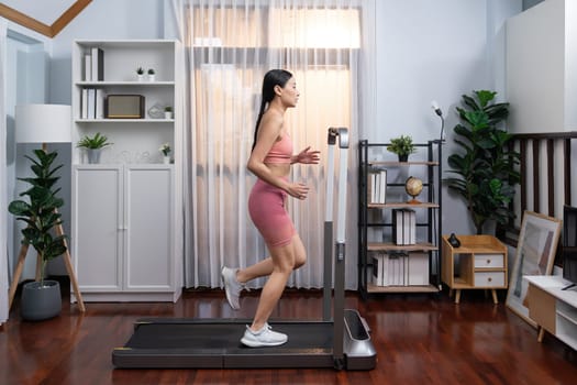 Full length side view of energetic and strong athletic asian woman running running machine at home. Pursuit of fit physique and commitment to healthy lifestyle with home workout and training. Vigorous