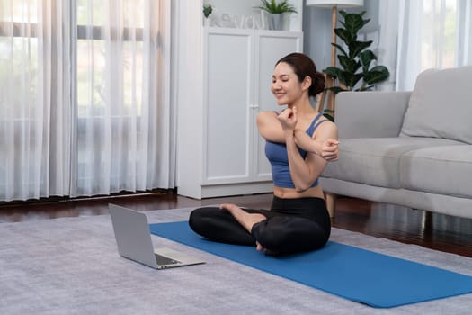 Asian woman in sportswear doing yoga exercise on fitness mat as her home workout training routine. Healthy body care lifestyle woman watching online yoga video on laptop. Vigorous